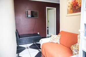 Une pièce comprenant un lit, un canapé noir, un téléviseur fixé au mur et une porte donnant sur une autre pièce.