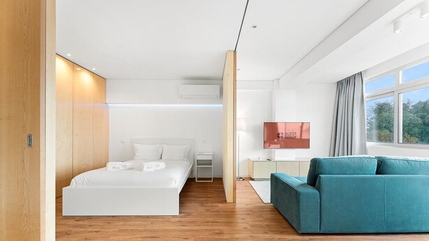 Une chambre d’hôtel moderne avec un grand lit, un canapé de couleur turquoise, une télévision et une fenêtre donnant sur des arbres.