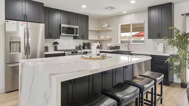 A modern kitchen with dark cabinets, a large island with stools, stainless steel appliances, and a tiled backsplash. - Desert Dream- Retreat with Heated Pool (Scottsdale)
