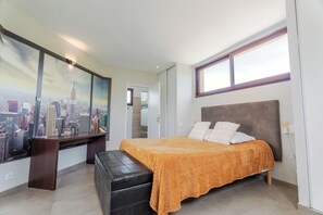 Une chambre avec un lit, un bureau, une grande fenêtre et une fresque murale représentant un paysage urbain.