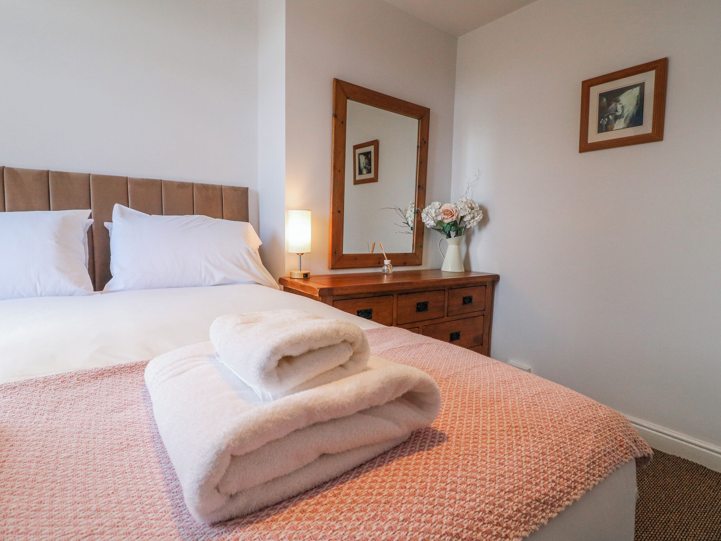 A neatly made bed with white linens and a folded towel, a bedside table with a lamp, a mirror, and a vase with flowers.