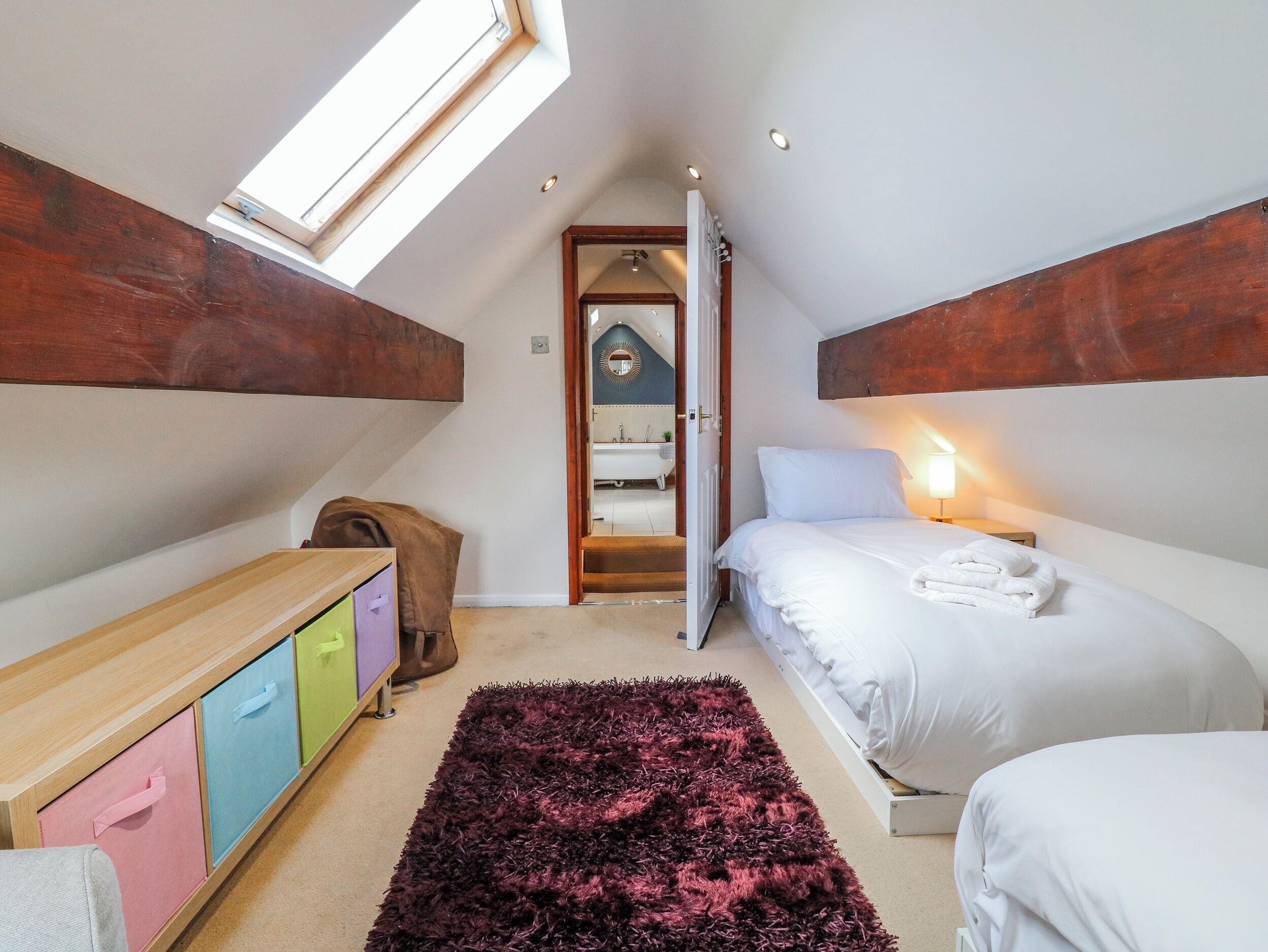 Quarto com claraboia, cama, mesa de cabeceira, guarda-roupa e banheiro visível por uma porta aberta.