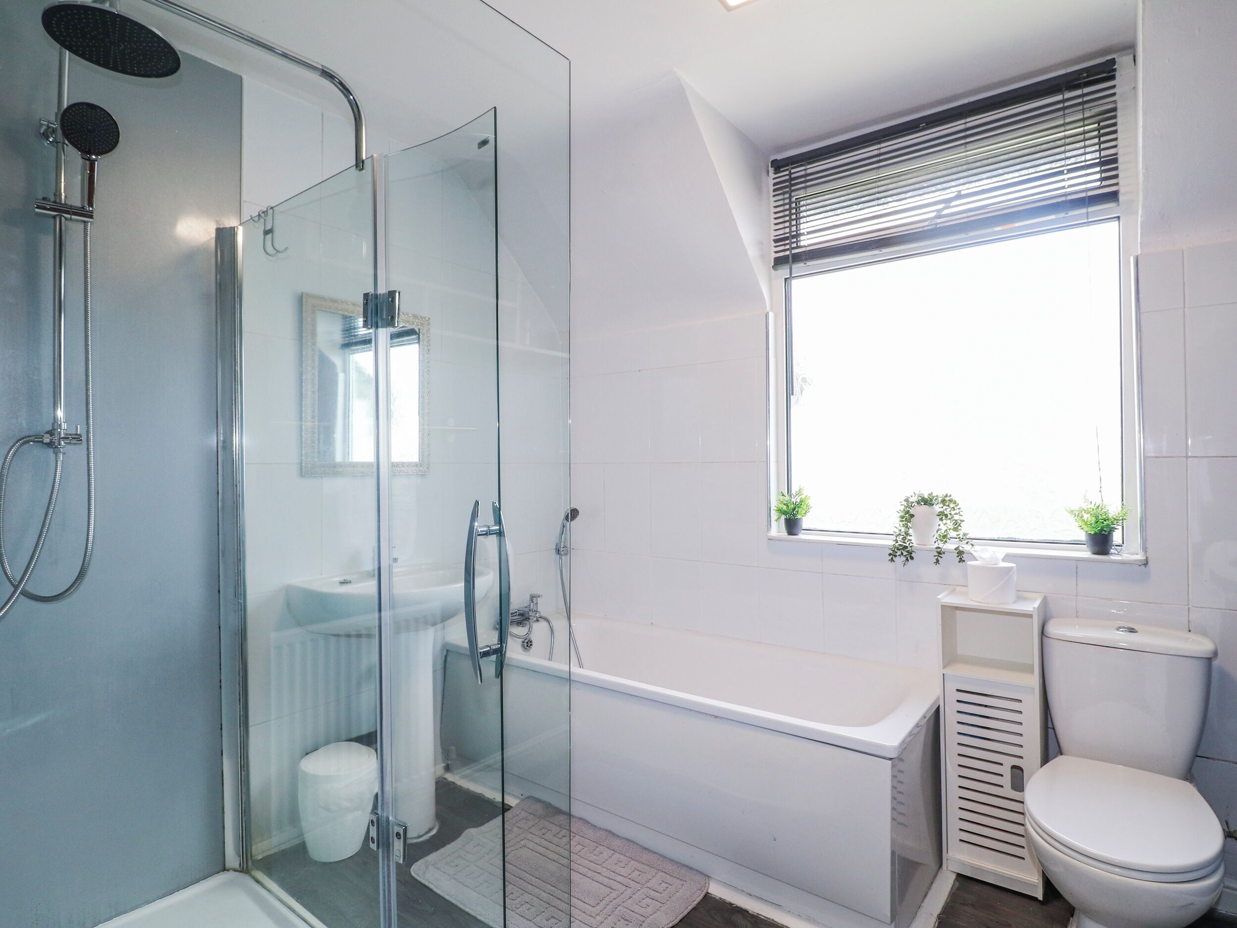 A modern bathroom with a glass-enclosed shower, a freestanding bathtub, a toilet, and a window with plants.