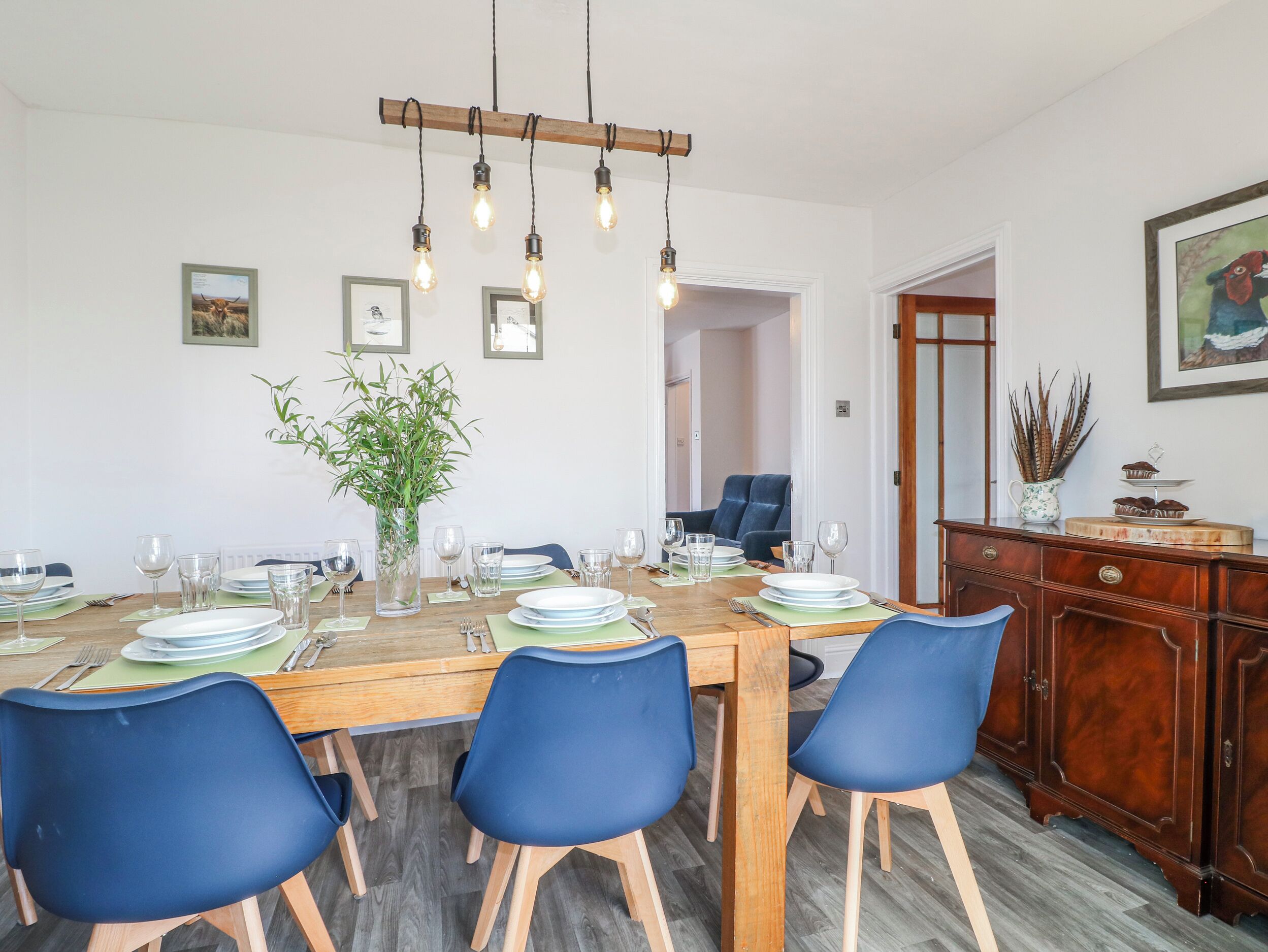 Un coin repas avec une table en bois dressée pour six personnes, des chaises bleues, un luminaire suspendu et des tableaux encadrés accrochés au mur.