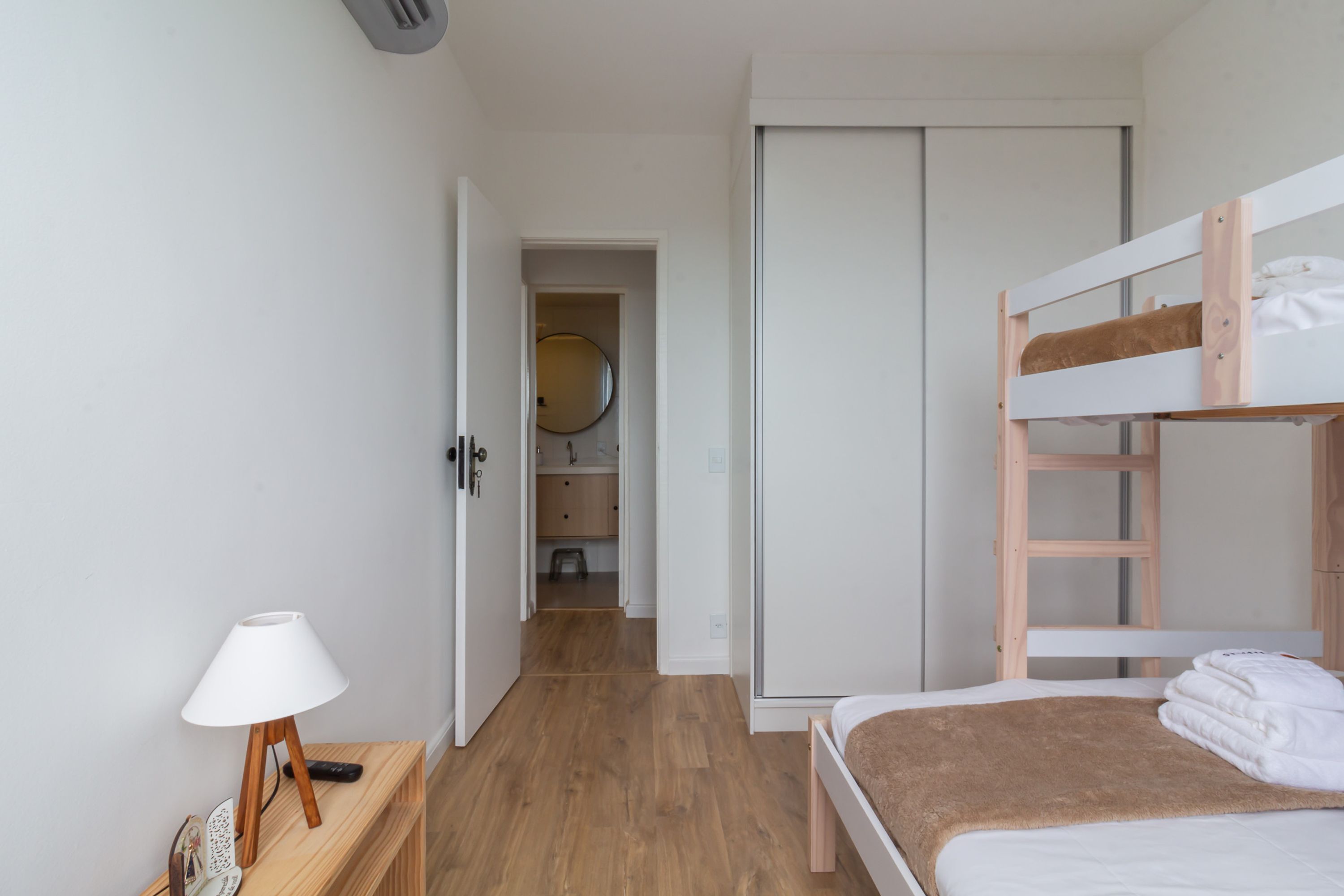 A bedroom with a bunk bed, a bedside table with a lamp, a wooden floor, and a wardrobe.