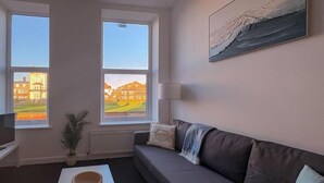 Un salon moderne avec un canapé gris, une table basse blanche et une vue sur les immeubles à travers de grandes fenêtres.