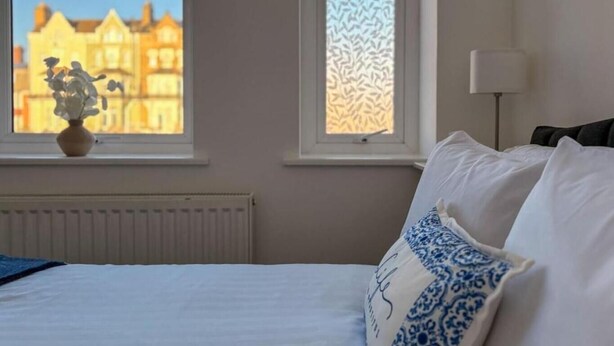 Une chambre d’hôtel avec un lit, une fenêtre donnant sur des immeubles, un radiateur et une lampe.