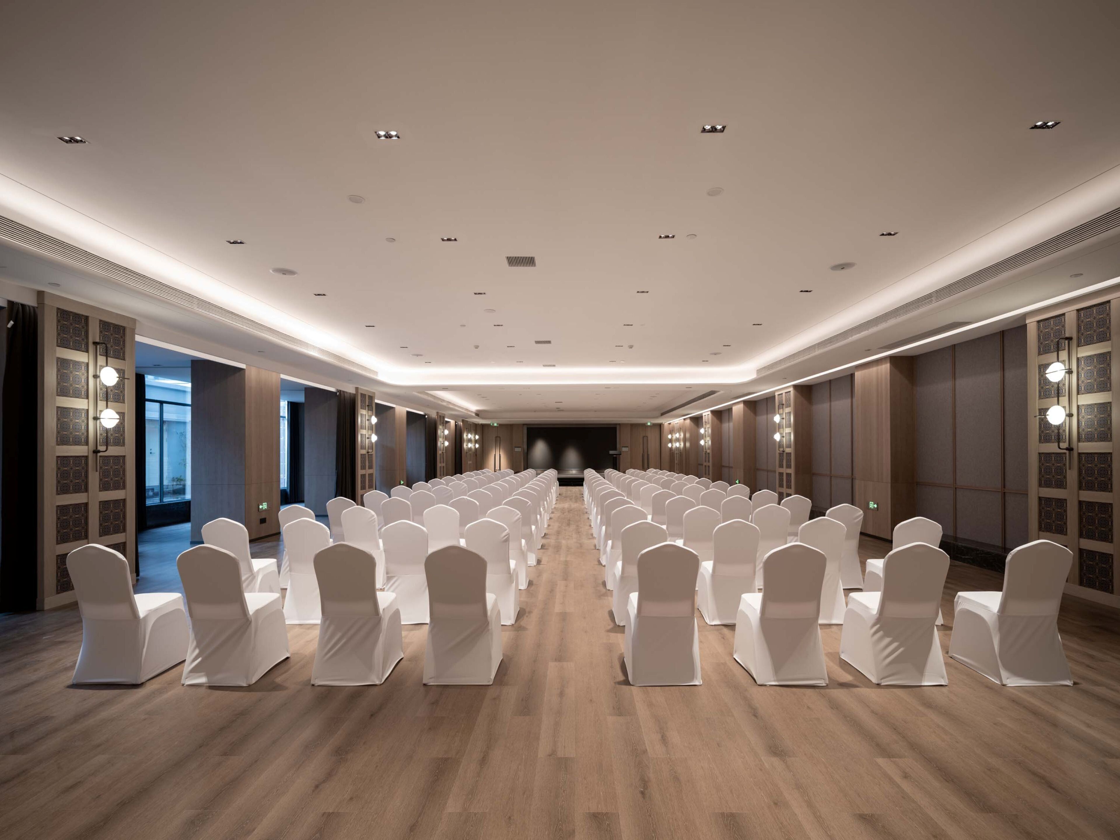 Un salón de conferencias con filas de sillas blancas, suelo de madera e iluminación empotrada.