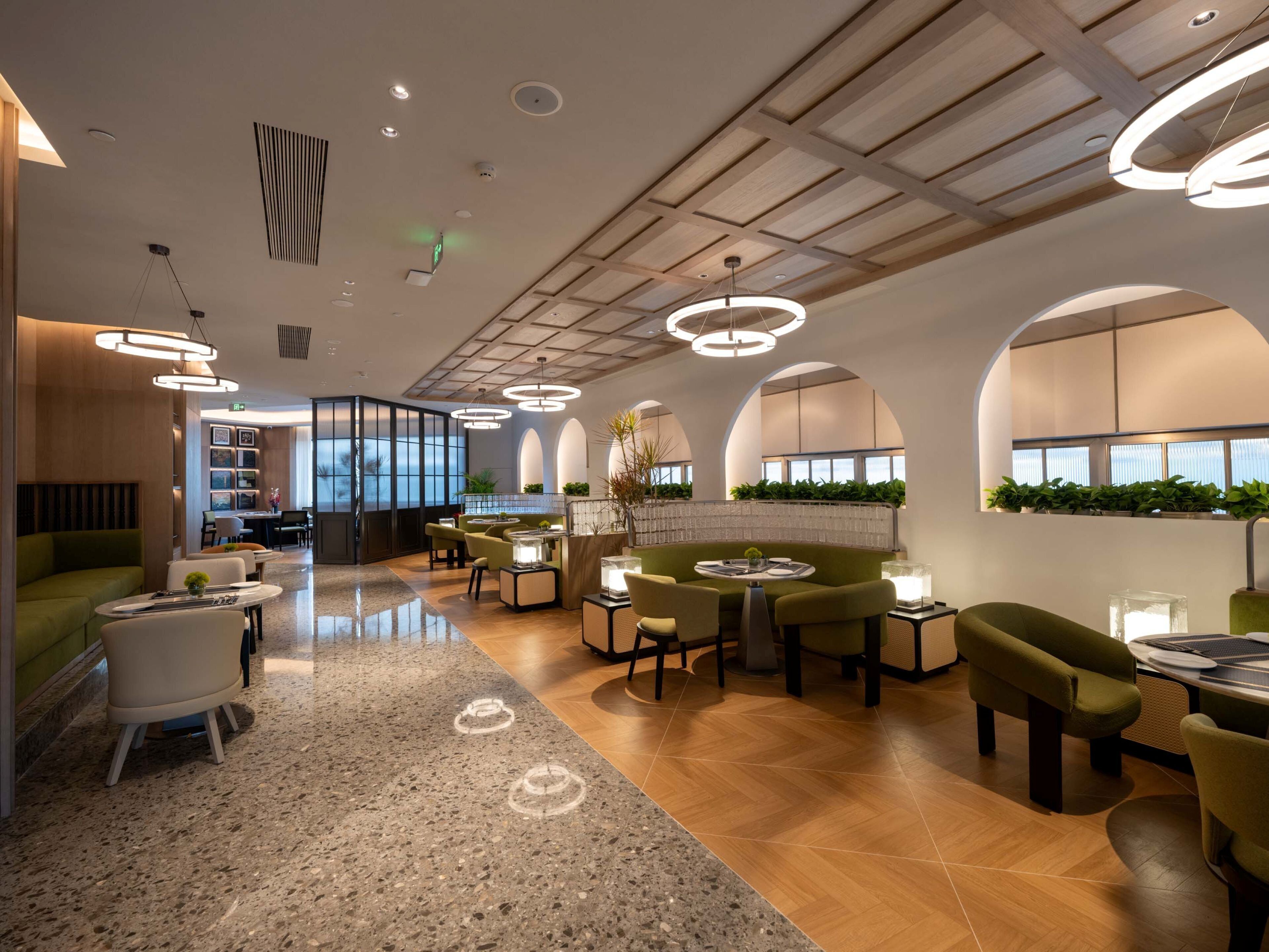 A modern interior with a mix of seating areas, including a green sofa and round tables, under a high ceiling with recessed lighting and a wooden panel design.