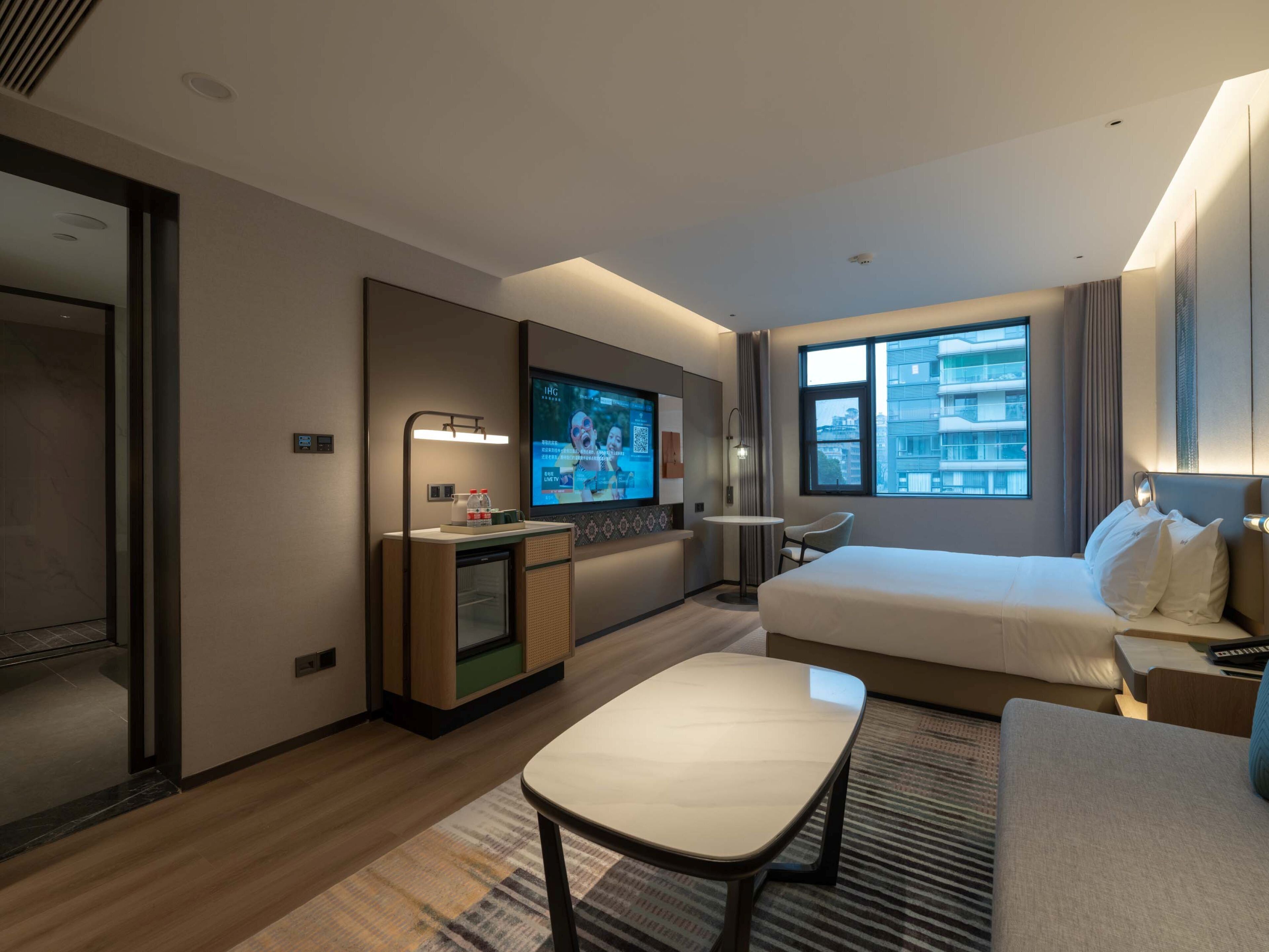 A hotel room with a bed, a desk, a TV, and a window with a city view.