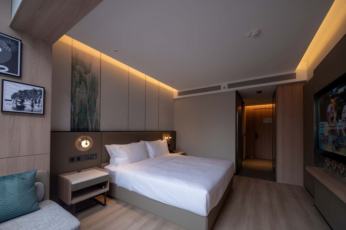 A modern hotel room with a large bed, a bedside table, a nightstand, and a seating area with a chair.