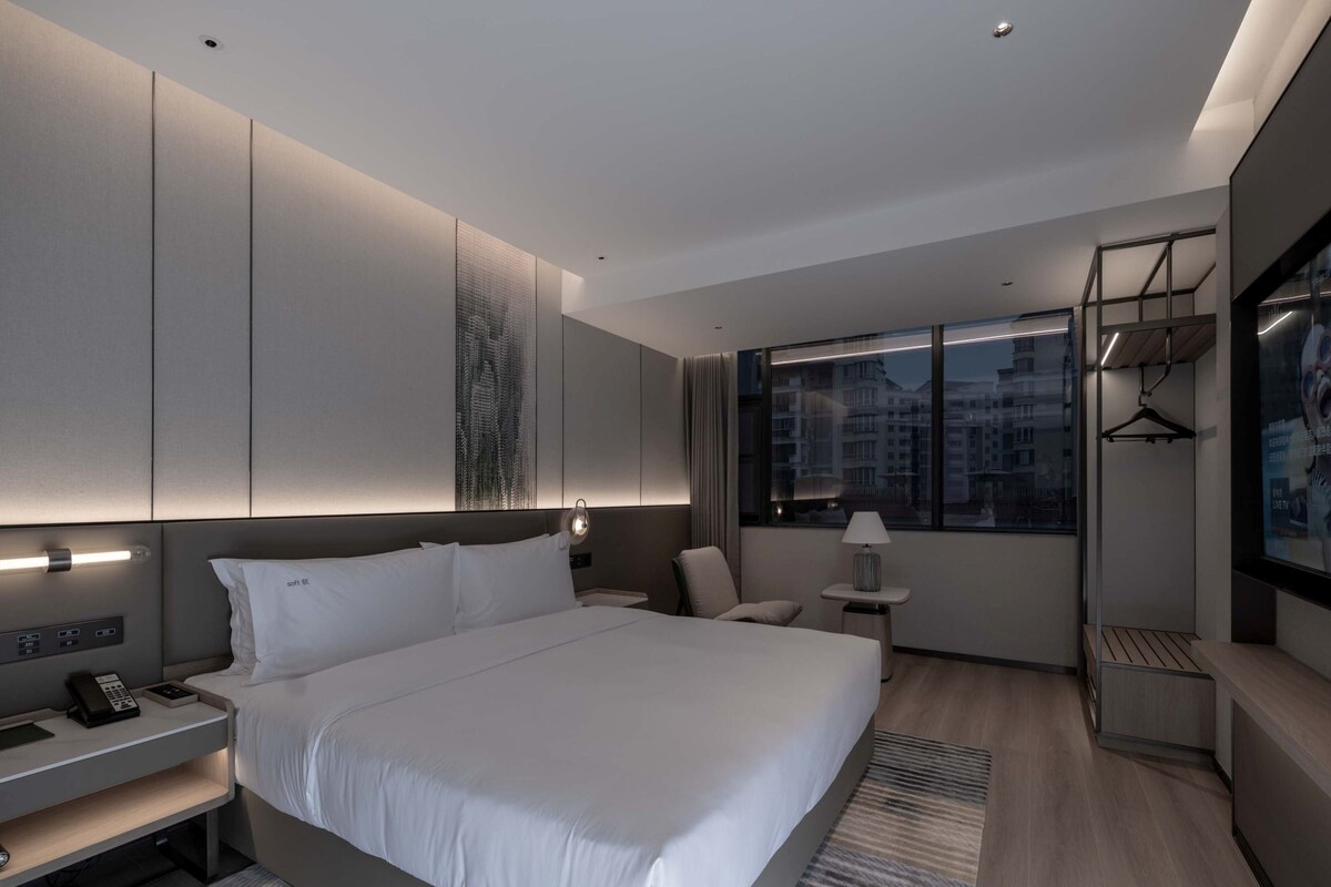 A modern hotel room with a large bed, bedside tables, a desk, a chair, a small table with a lamp, and a view of the city through a large window.