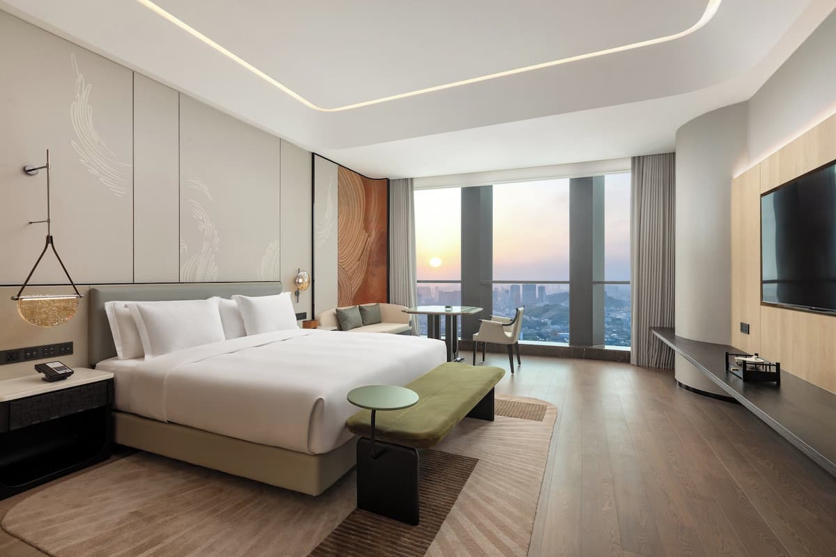 Ein modernes Hotelzimmer mit einem großen Bett, einem kleinen Tisch, einer Bank, einem Fernseher und Blick auf die Stadt.