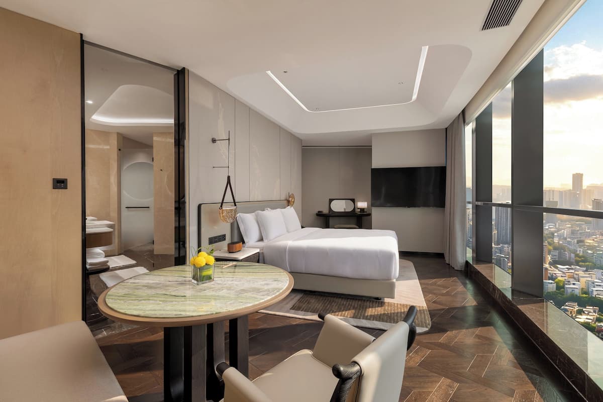Ein modernes Hotelzimmer mit einem großen Bett, einem runden Tisch mit einer Vase, Blick auf die Stadt und einem Balkon.