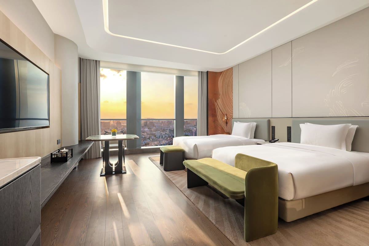 Ein modernes Hotelzimmer mit einem großen Bett, einem Schreibtisch, einem Sessel, einem Fernseher und Blick auf die Stadt.