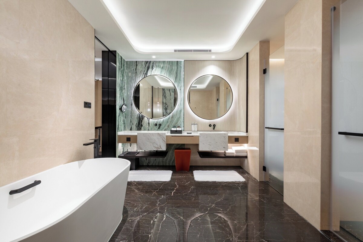 A modern bathroom with a freestanding bathtub, two sinks with round mirrors, and a marble backsplash.