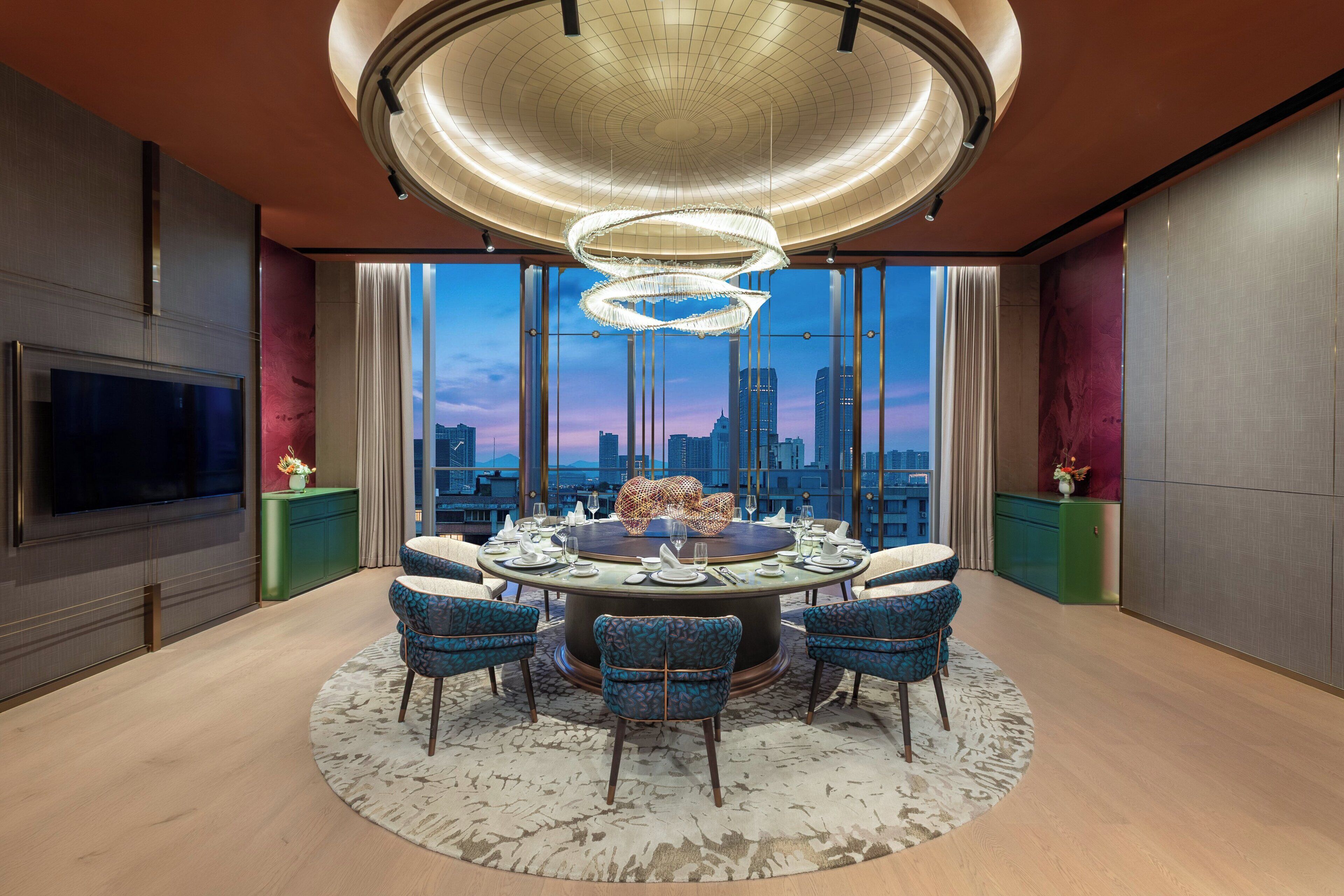A modern dining room with a round table, chairs, a chandelier, and a city view.