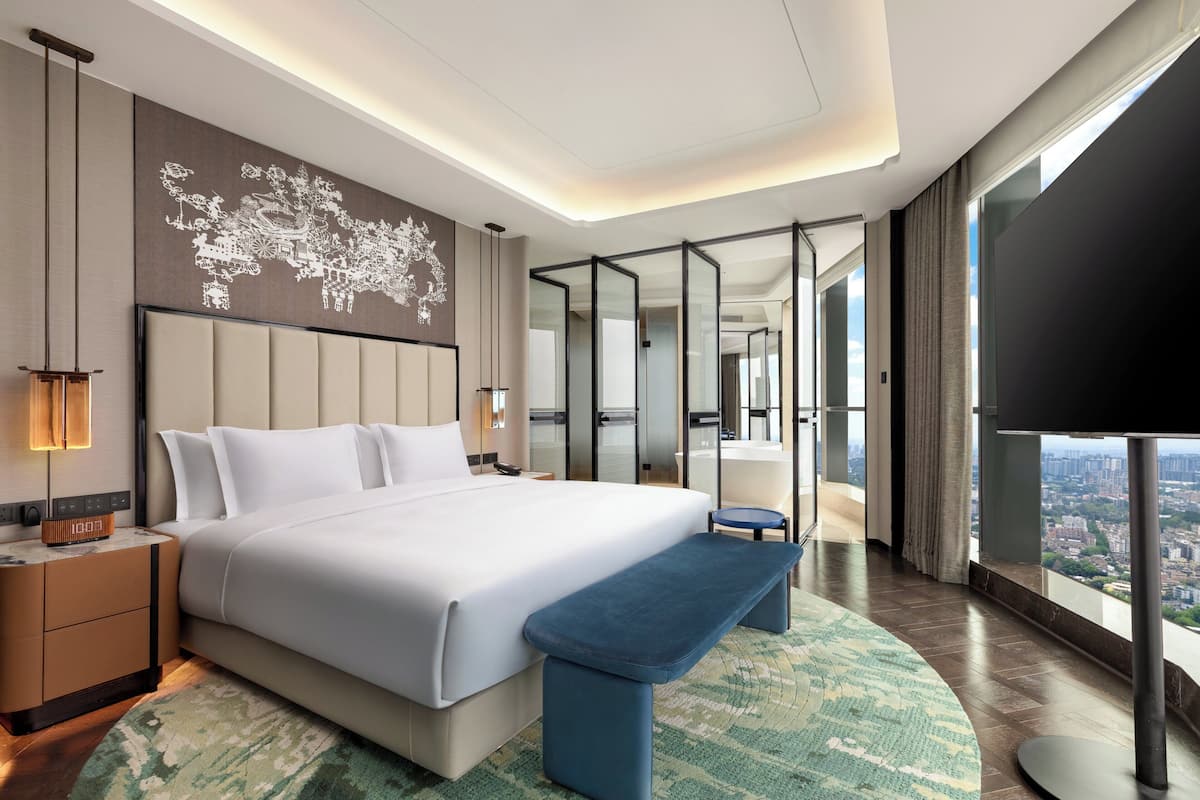 Ein modernes Hotelzimmer mit einem großen Bett, einer Bank, einem Wandbild mit einer Weltkarte, einem Fernseher und Blick auf die Stadt.