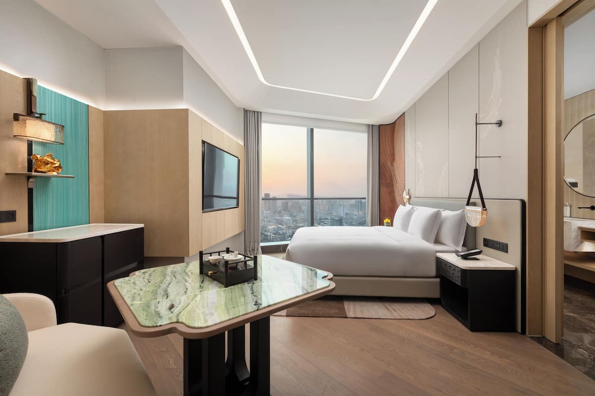 Ein modernes Hotelzimmer mit einem großen Bett, einem Schreibtisch mit Marmorplatte, einem Fernseher und Blick auf die Stadt.