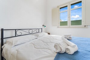 Un lit avec une literie blanche, une couverture bleue et des serviettes enroulées. Il y a une fenêtre d’où l’on peut voir le ciel et un bâtiment.