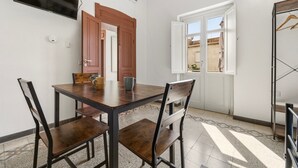 Uma área de refeições com mesa e cadeiras de madeira, uma televisão fixada na parede e uma janela com porta aberta.
