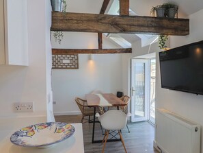A modern kitchen with a wooden beam ceiling, a dining area with wicker chairs, a large TV, and a view through a glass door. - Avocet (Lyme Regis)