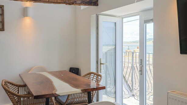 Una habitación con una mesa de madera, sillas de mimbre, un ventanal de vidrio que da a un balcón con vistas al mar.