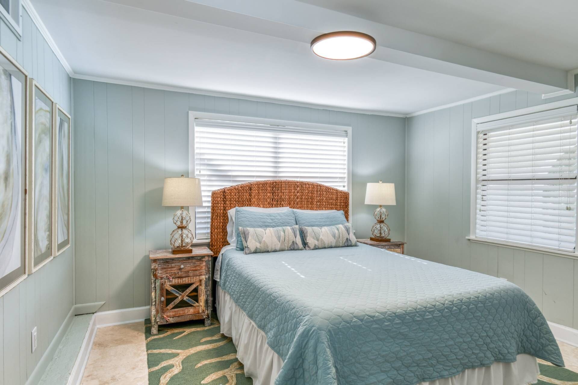 A bedroom with a bed, two bedside tables, a headboard, and two windows with blinds.