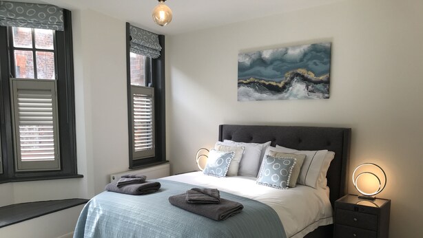 A bedroom with a bed, bedside tables, a window with blinds, a wall-mounted artwork, and a radiator. - Boutique Style 2 bed Maisonette (Watlington)
