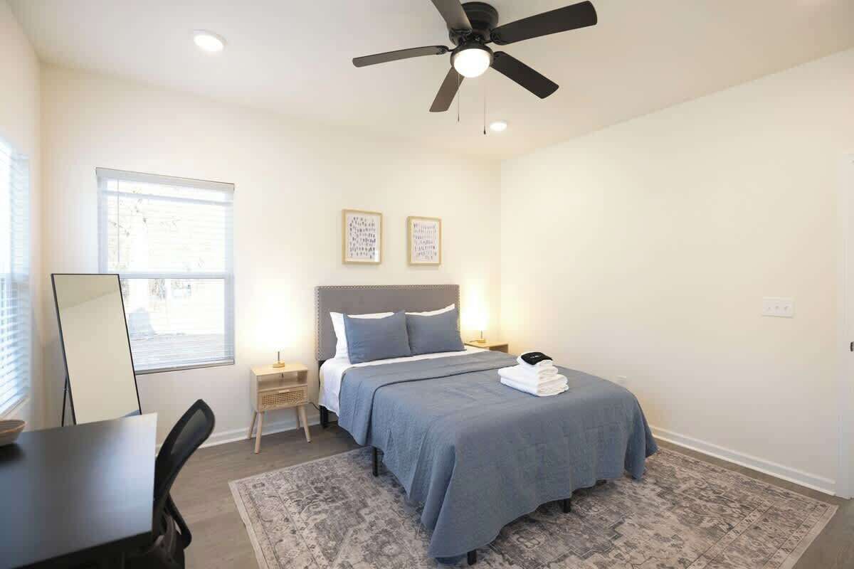 A bedroom with a bed, a desk, a chair, a ceiling fan, and a window.