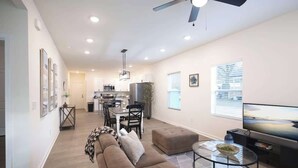A modern living room with a large sofa, a coffee table, and a television. There is a dining area with a table and chairs, and a kitchen in the background. - 5 Min to Elon | 7 Min to Downtown | Washer + Dryer (Burlington)