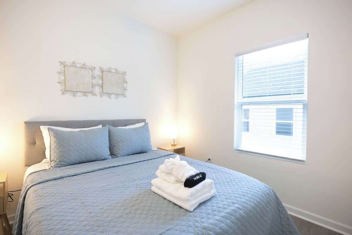 A neatly made bed with a quilted bedspread and pillows, a bedside lamp, and a window with blinds.