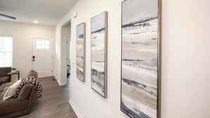 A hallway with three abstract paintings on the wall, a brown couch, and a door at the end. - 5 Min to Elon | 7 Min to Downtown | Washer + Dryer (Burlington)