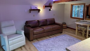 Una sala de estar acogedora con un sofá de cuero marrón, un sillón blanco, una mesa de comedor de madera con sillas y un ventanal con vistas a árboles.