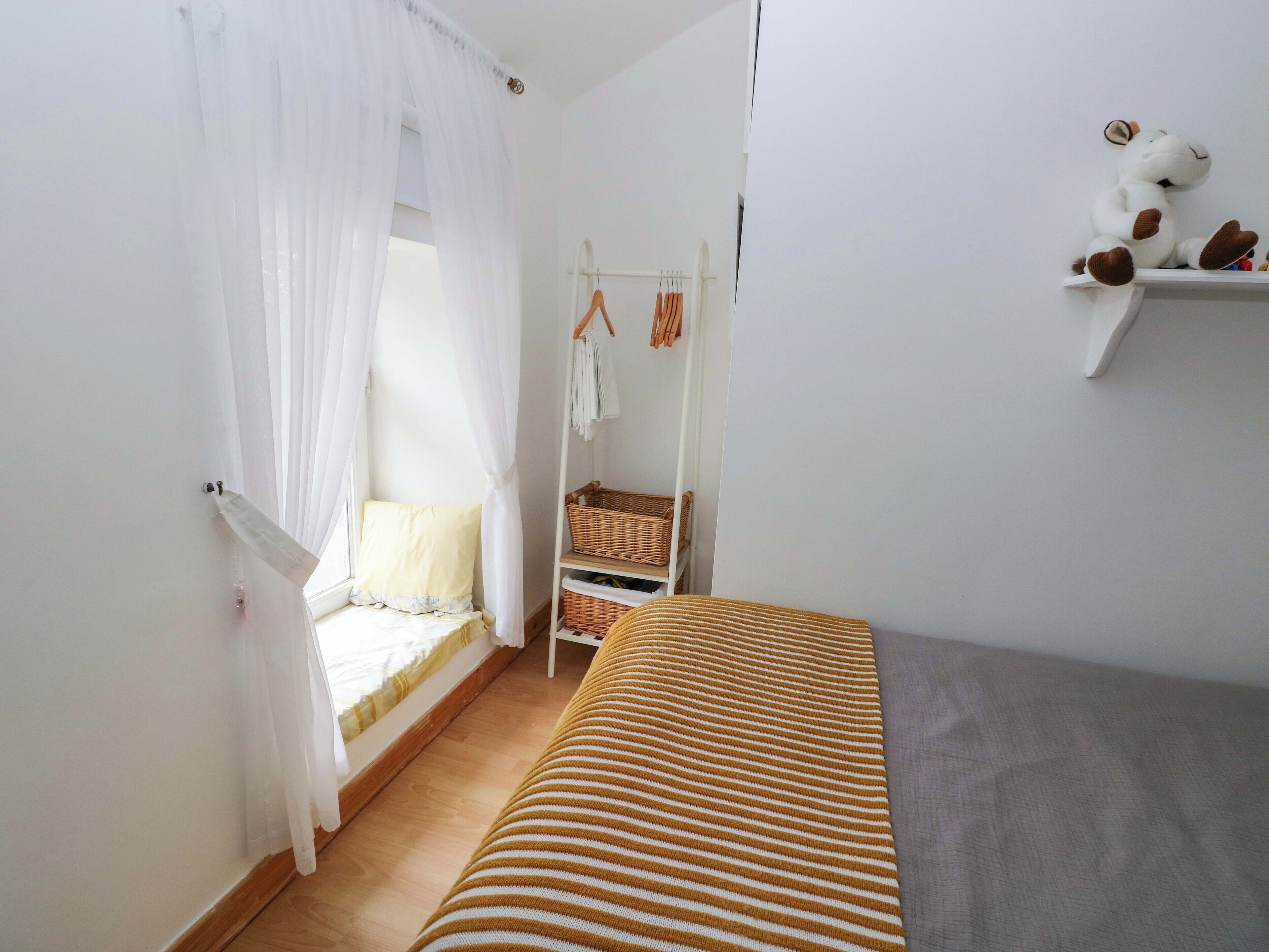 A bedroom with a bed, a window with sheer curtains, a bedside table, a hanging rack with clothes, and a shelf with stuffed toys.