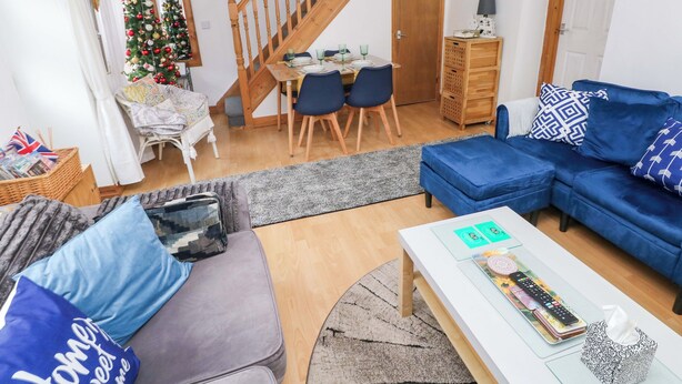 Un salon chaleureux avec un canapé bleu, un divan gris et une table basse blanche. Un escalier en bois mène à une autre pièce, on y trouve un coin repas avec des chaises et un sapin de Noël.