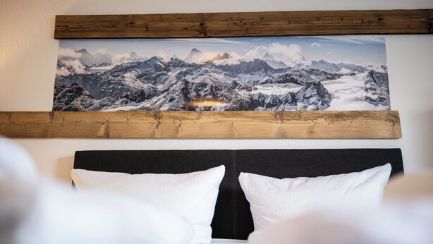 Een slaapkamer met een berglandschap schilderij boven het bed, een zwart hoofdeinde en witte kussens.