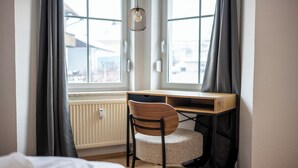 Een slaapkamer met een bed, een bureau en een stoel. Er zijn twee ramen met gordijnen, een radiator en een hanglamp.