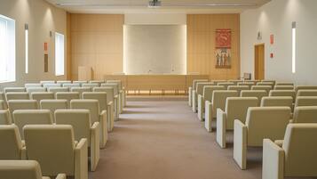 Un salón de conferencias con filas de sillas color beige, un atril al frente y un ventanal grande a la izquierda.