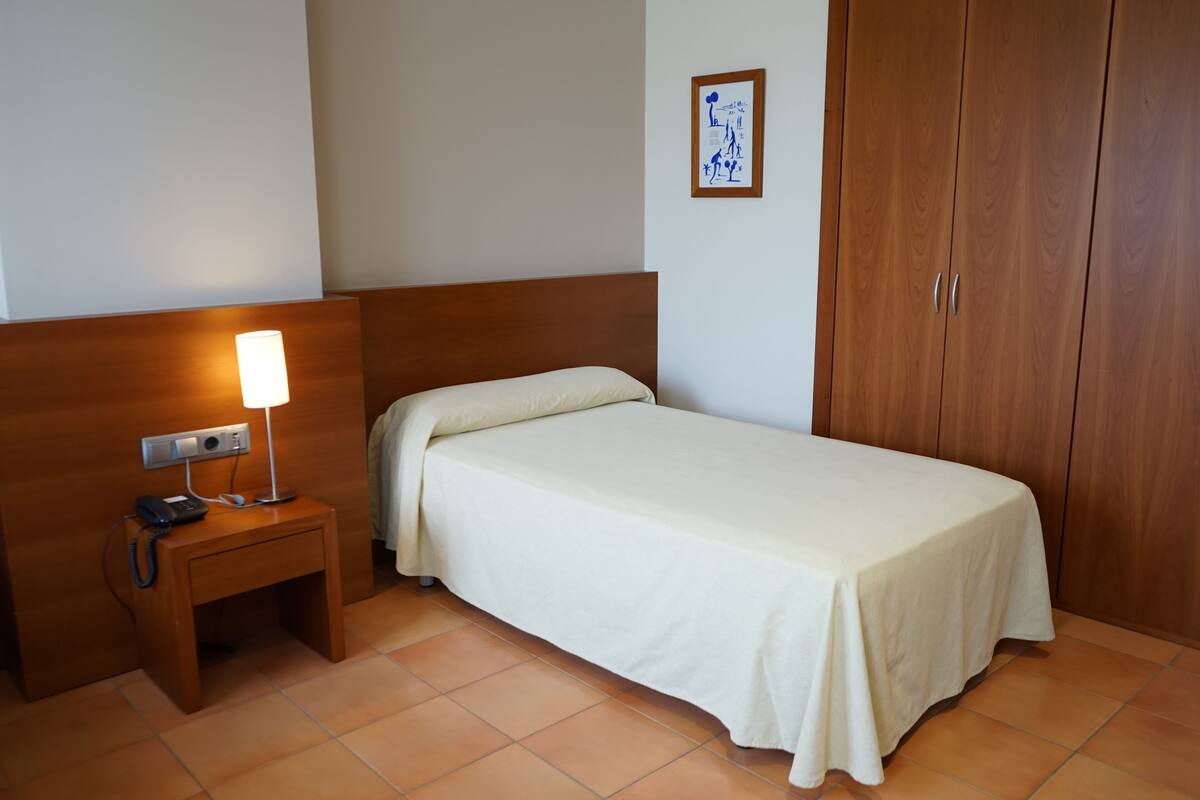 Een hotelkamer met een enkel bed, een nachtkastje met een lamp, een telefoon en een kledingkast.