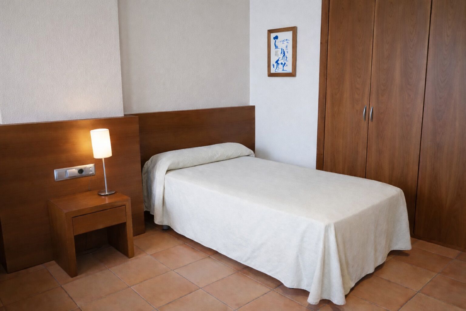 A single bed with a white bedspread, a bedside table with a lamp, and a wooden wardrobe.