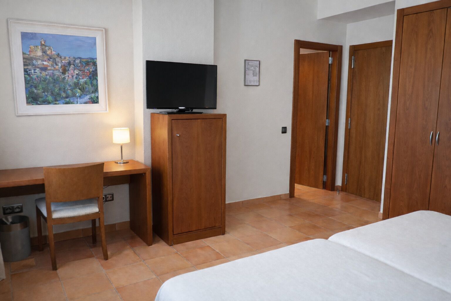 A hotel room with a bed, a desk with a chair, a television, and wooden wardrobes.