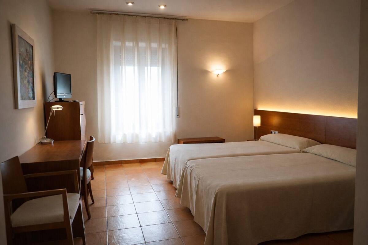Een hotelkamer met een bureau, twee bedden, een televisie en een raam met gordijnen.