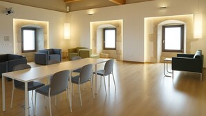 A spacious room with a long table and chairs, a sofa, and a small table with chairs. There are large windows with arched tops and wall-mounted lighting. - Hotel Santuari Balaguer (Balaguer)