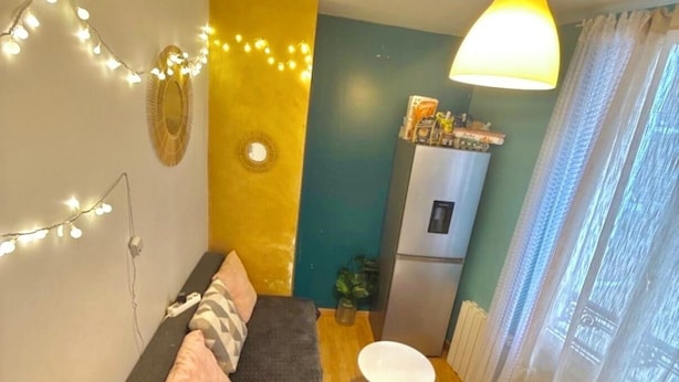 Un espacio habitable pequeño con un sofá, dos mesitas, un refrigerador y luces de cadena.