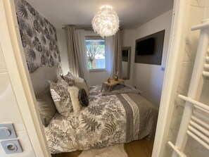 Un dormitorio con cama, una mesita de noche, televisor en la pared y ventana con cortinas.