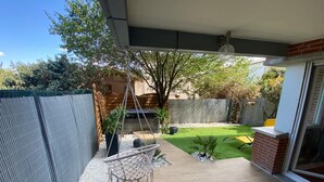 A patio with a hanging chair, a small table with a bowl, and a view of a backyard with a lawn and trees. - Le Jungle - 2p - Blagnac (Blagnac)