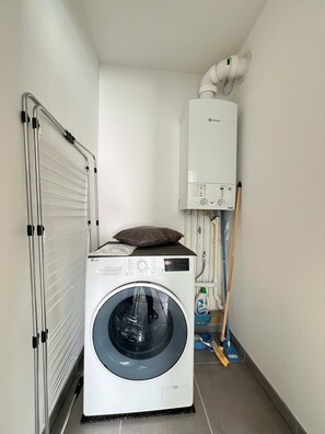 A compact laundry room with a front-loading washing machine, a drying rack, a heater, and cleaning supplies. - Le Lamartine - 4p - Bords de Lac - Parking (Frouzins)