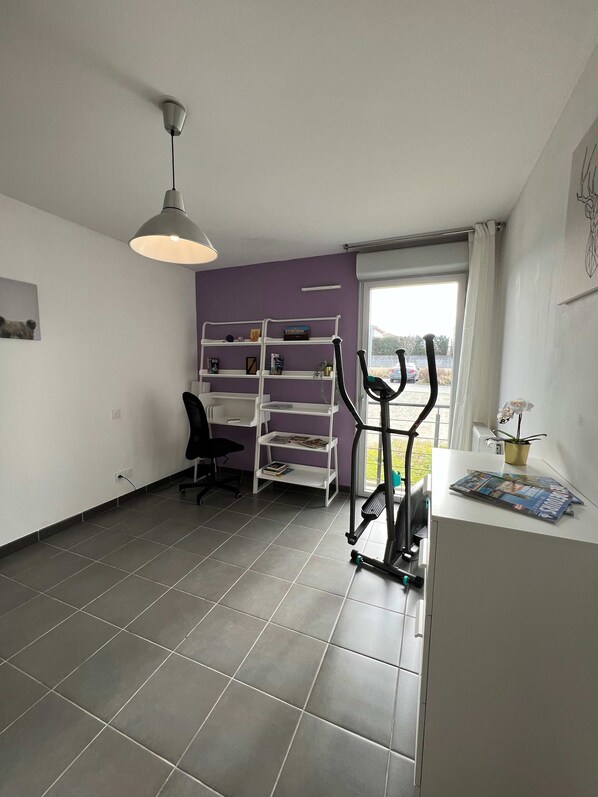A room with a grey tiled floor, a white desk with a chair, a standing desk with shelves, a treadmill, and a window with a view of the outside. - Le Quinze Sols - 5p - Blagnac - Parking (Blagnac)