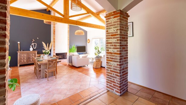A spacious living area with a high ceiling, wooden beams, a brick fireplace, and a dining area with a table and chairs. - La Tendresse - 10p - Piscine - Jardin (Mondonville)